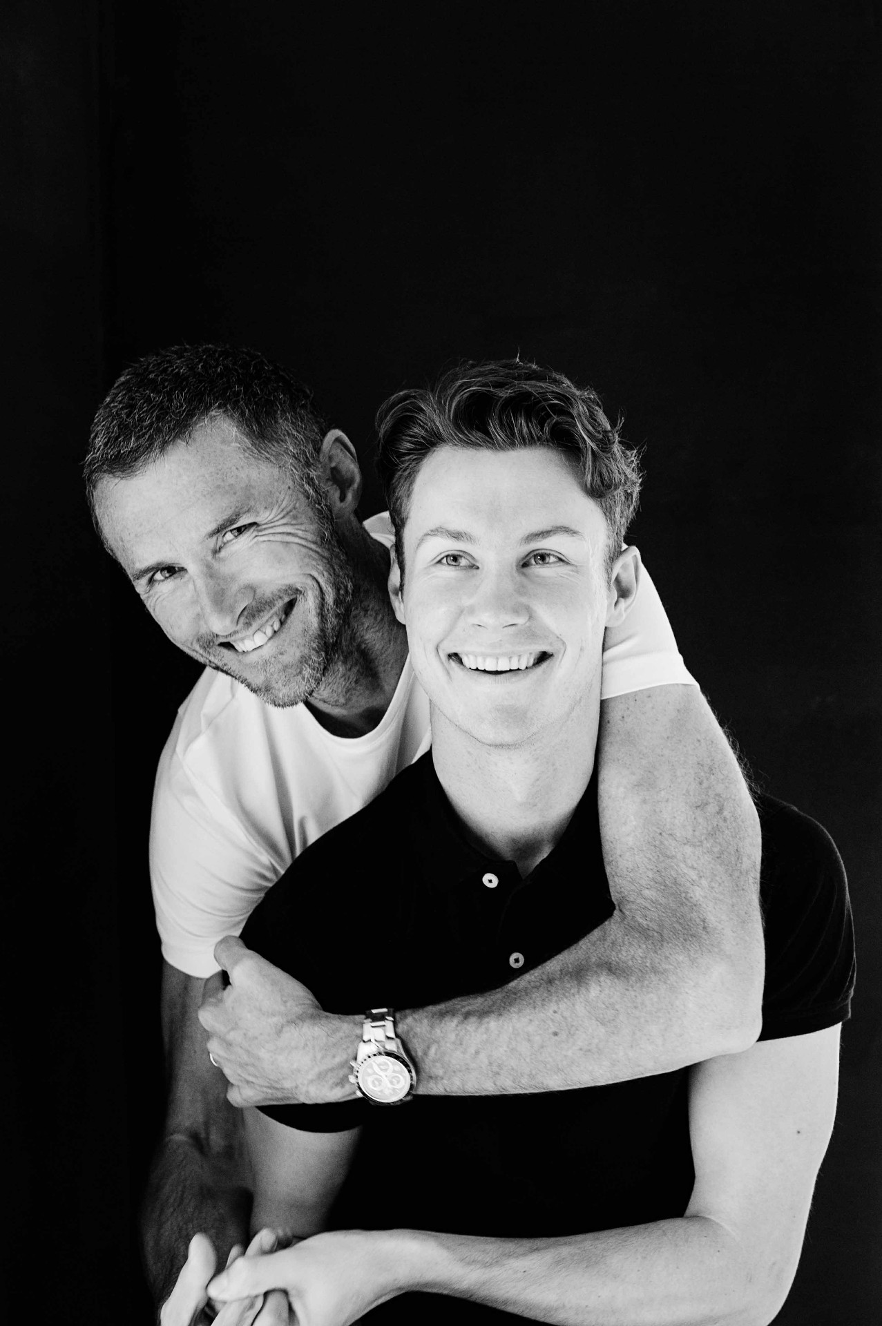 A father wrapping his arm around his grown son, both laughing.