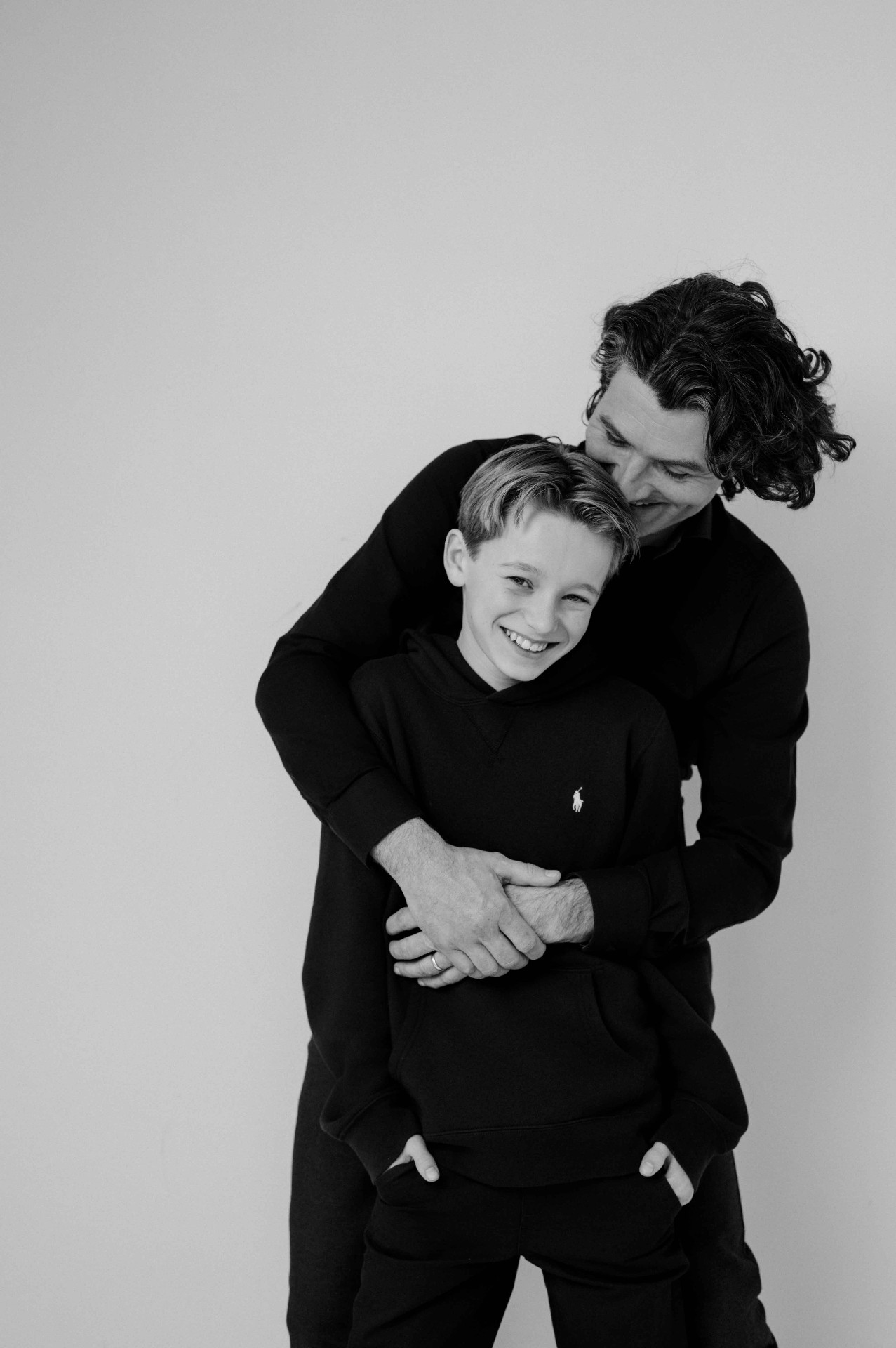 A father hugging his young son from behind, both in black, soft black and white.