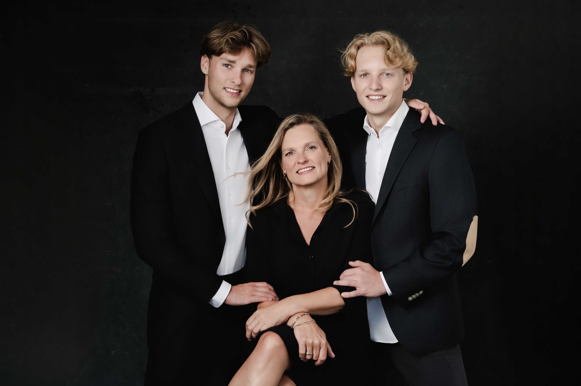 A mother seated between her two grown sons in elegant black tailoring.